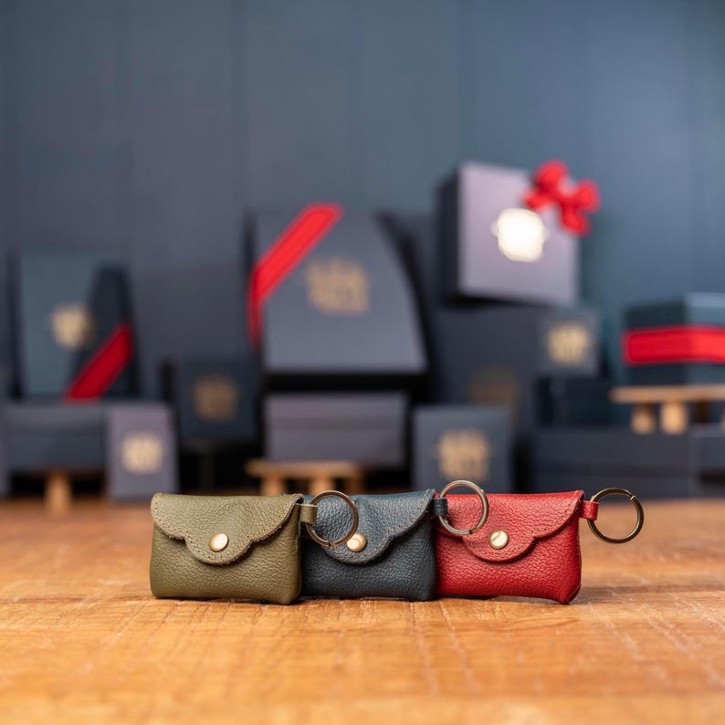 May include: Three small envelope-style pouches made of textured leather, each featuring a gold-tone snap button closure and a metal keyring attached to the side. The pouches are colored olive green, dark gray, and bright red, arranged in a row.