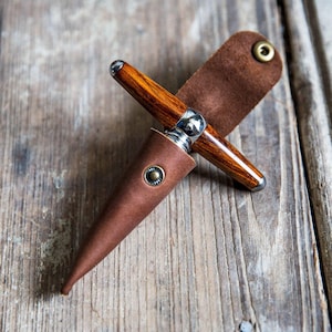 May include: A handcrafted wooden pen with a brown leather sheath. The pen features a polished wood grain barrel with silver-toned metal accents. The sheath is a rich brown color with a snap closure and a loop for carrying.