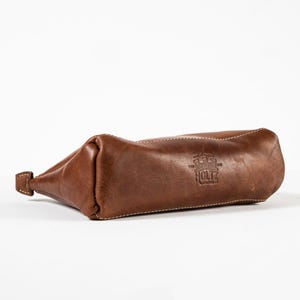 May include: A brown leather pouch with a rectangular base and a zippered closure. The leather features visible stitching along the edges and a stamped logo on the side that reads Holtz Leather Co. with a castle icon above the text.