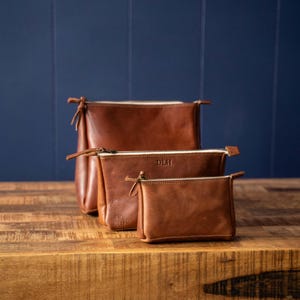 May include: Three brown leather pouches of varying sizes arranged in a row. Each pouch features a top zipper closure and visible stitching. The middle pouch is embossed with the letters DLH, and the smallest pouch has a small logo stamp.