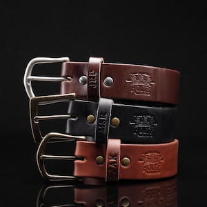 May include: Three stacked leather belts in dark brown, black, and tan. Each belt features a metal buckle, a leather keeper loop embossed with initials JRP, MSR, or JAK, and a stamped logo reading HOLTZ LEATHER CO with a crown icon.