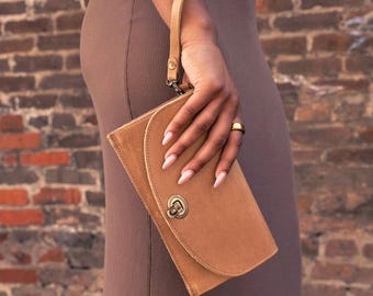 May include: A tan leather wristlet clutch featuring a curved front flap, a brass-toned metal turn-lock closure, and a matching leather wrist strap. The accessory has a smooth, matte finish and visible stitching along the edges of the flap.