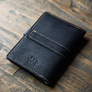 May include: A black leather wallet featuring a horizontal strap closure with visible light-colored stitching along the edges. The front of the wallet is embossed with a circular logo that includes the text Holtz Leather Co.