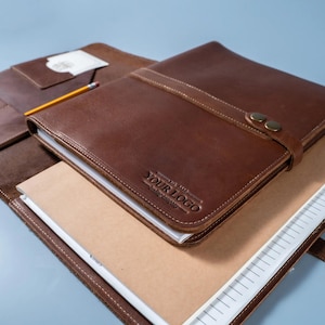 May include: A brown leather portfolio folder featuring a strap with two metal snap buttons, embossed with the text YOUR LOGO, containing a notebook with lined paper and a yellow pencil resting on the interior flap of the leather cover.