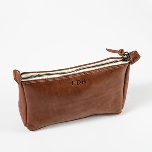 May include: A brown leather zippered pouch featuring the embossed letters CDH on the front. The rectangular accessory has a top zipper closure with a leather pull tab and visible stitching along the edges, revealing a light-colored interior lining.