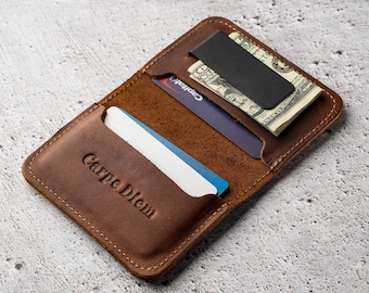 Puede incluir: Una cartera de cuero marrón con un clip de metal negro mate para sujetar billetes. La cartera tiene una ranura con una tarjeta blanca que lleva grabado el texto Carpe Diem, y una segunda ranura que contiene una tarjeta de color azul oscuro.