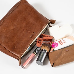 May include: A brown leather zippered pouch with the initials CDH embossed on the front. The pouch is open, revealing a collection of items including a pink lip gloss, an EcoTools makeup brush, a terracotta hair clip, a white sponge, and a tube of EOS Shea Better hand cream, alongside a small brown wallet.