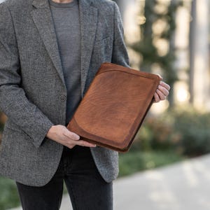 May include: A brown leather portfolio with a textured surface. The portfolio has a stitched border and a rounded top corner. The person is wearing a gray blazer and dark jeans.