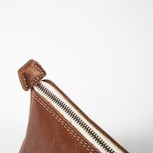 May include: A brown leather pouch featuring a white zipper closure with a matching brown leather pull tab. The pouch is constructed with visible light-colored stitching along the seams, highlighting the texture and edge of the leather material.