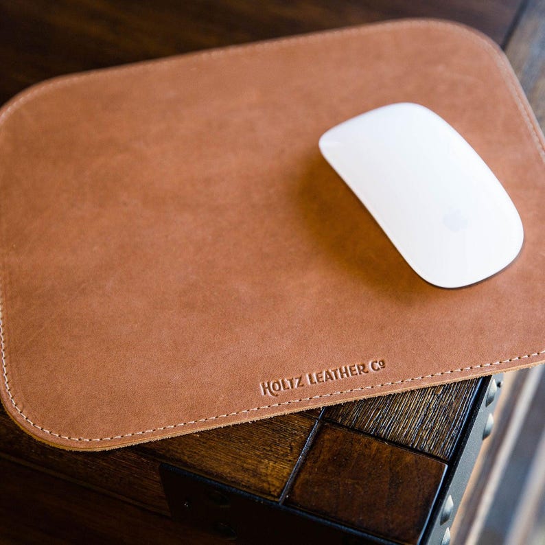 May include: A rectangular tan leather mouse pad with rounded corners and visible stitching along the edges. The text HOLTZ LEATHER CO is embossed in the bottom center. A smooth white wireless computer mouse rests on the surface of the pad.