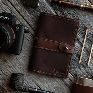 May include: A dark brown leather notebook cover featuring a horizontal strap with a brass stud closure and the embossed name J. Muir in the bottom right corner. The cover is stitched with light brown thread along the edges and the strap.