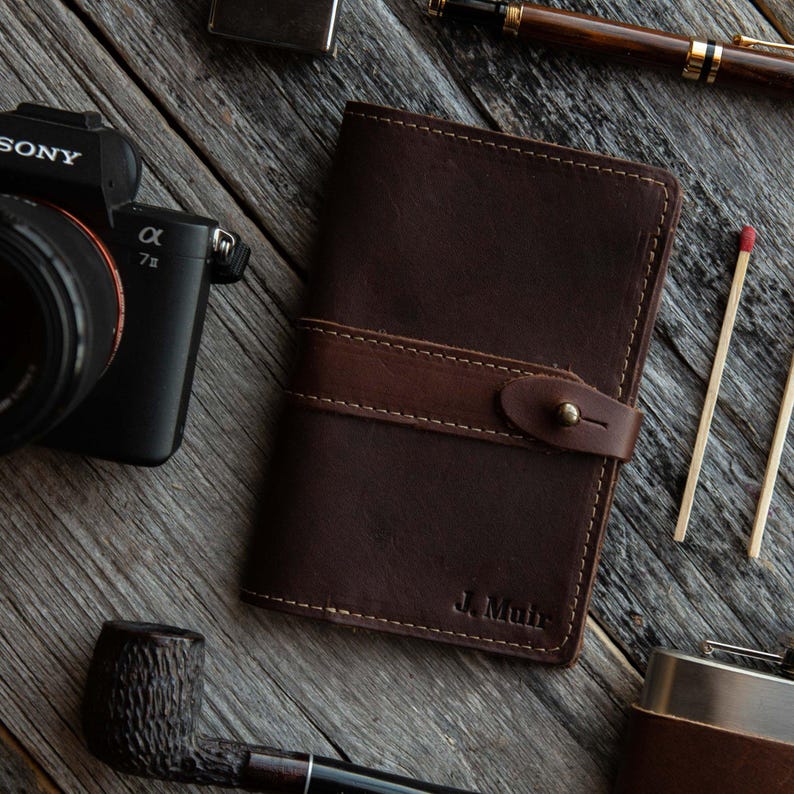 May include: A dark brown leather notebook cover featuring a horizontal strap with a brass stud closure and the embossed name J. Muir in the bottom right corner. The cover is stitched with light brown thread along the edges and the strap.