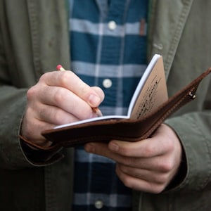 May include: A brown leather notebook cover holding a small booklet with the text "FIELD NOTES BRAND BOOK" printed on the cover. A yellow pencil is held against the white paper pages of the open notebook, which is worn by a person in a plaid shirt.