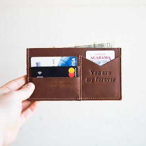 May include: A brown leather card holder wallet with visible stitching, featuring multiple slots containing credit cards, an Alabama driver license, and a twenty-dollar bill. The leather is embossed with the text You are my forever.