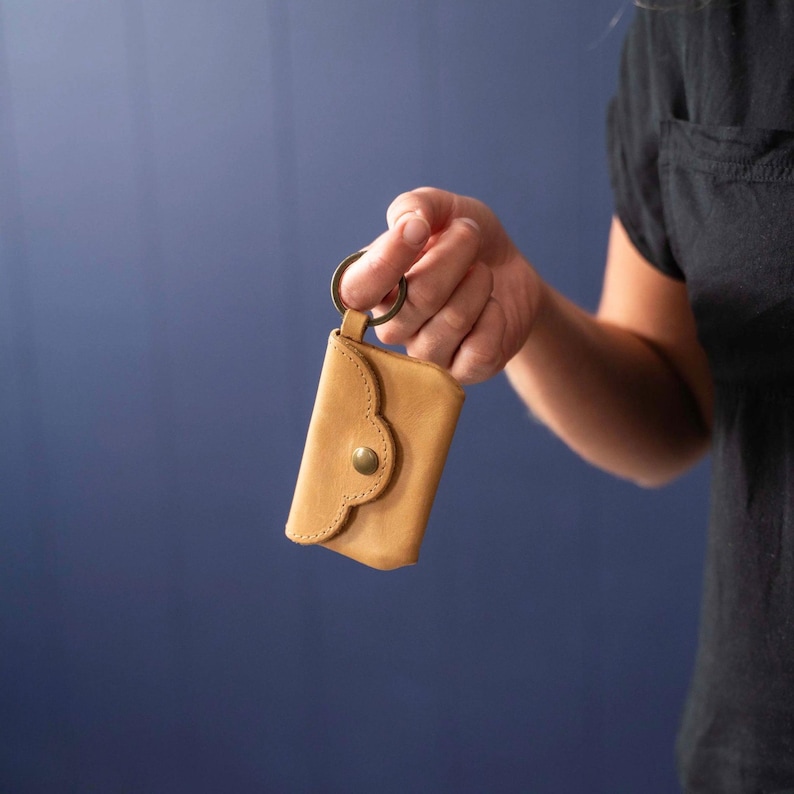 May include: A tan leather key pouch featuring a scalloped flap closure secured with a single brass-colored metal snap button. The pouch is attached to a circular metal key ring, providing a compact storage solution for keys or small items.