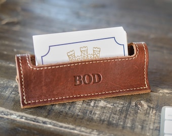 May include: A brown leather business card holder with visible white stitching along the edges. The front of the holder is embossed with the letters BOD. A stack of white business cards with a blue border and a gold castle graphic is inside.