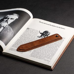 May include: A brown leather bookmark with a pointed end and a metal snap fastener, resting on an open book. The book page features the text The men who turned the longhorn into big business and a black and white illustration of a longhorn steer.