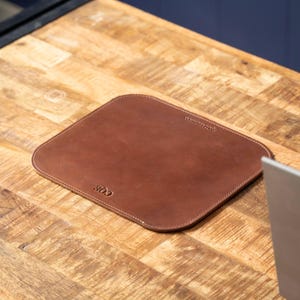 May include: A rectangular brown leather mouse pad with rounded corners and stitched edges. The surface features the embossed letters CQE in the bottom left corner and a small brand logo embossed in the top right corner of the leather mat.