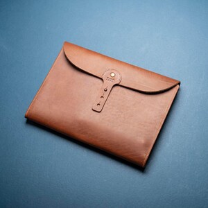 May include: A tan leather document folder featuring a curved flap closure secured by a vertical strap with a brass-colored metal stud. The strap is embossed with the name LEO and includes three additional circular cutouts for adjustment.