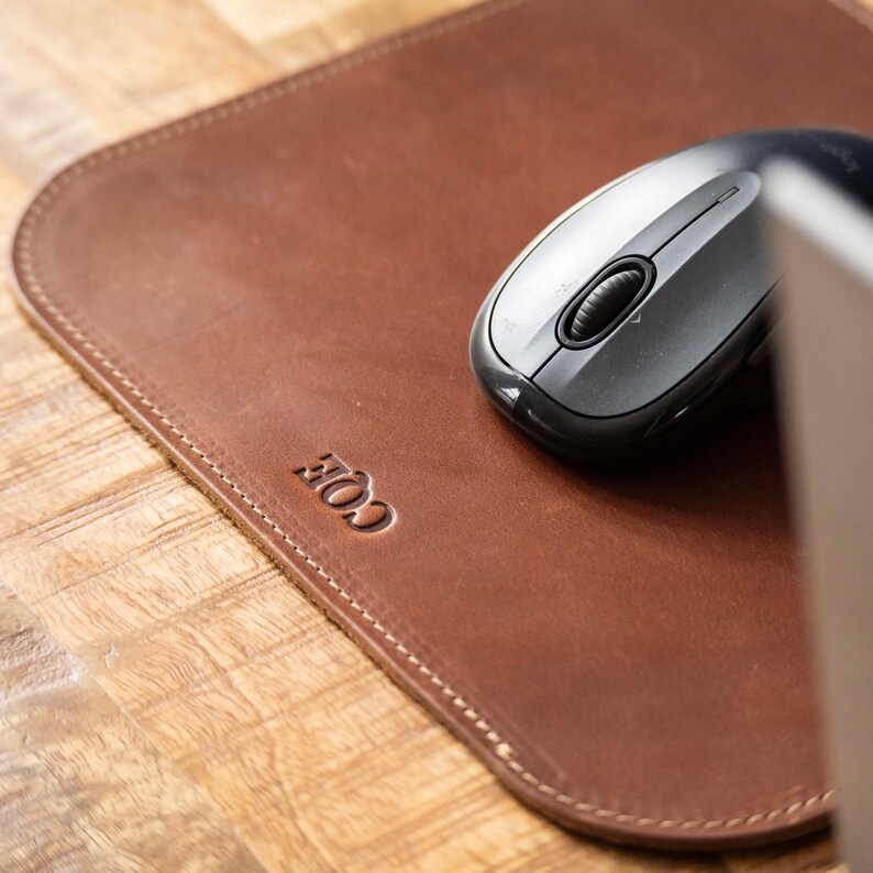 May include: A smooth brown leather mouse pad with rounded corners and stitched edges. The word COE is embossed in the lower left corner. A dark gray wireless computer mouse with a central scroll wheel rests on the surface of the leather pad.