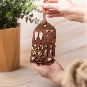 May include: A brown leather earring holder with a loop at the top. The holder has six slots for earrings and a ring holder. The holder is filled with various earrings, including gold hoops, teardrop-shaped earrings with gold accents, and white ceramic earrings with gold flecks.