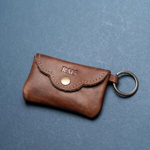 May include: A brown leather key pouch featuring a scalloped flap, a gold-toned metal snap closure, and a circular metal keyring attached to the side. The letters RMS are embossed in a serif font on the front flap of the leather accessory.