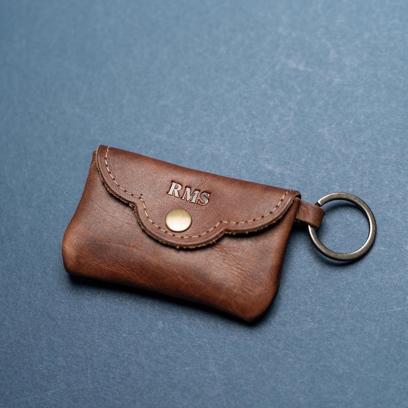 May include: A brown leather key pouch featuring a scalloped flap, a gold-toned metal snap closure, and a circular metal keyring attached to the side. The letters RMS are embossed in a serif font on the front flap of the leather accessory.