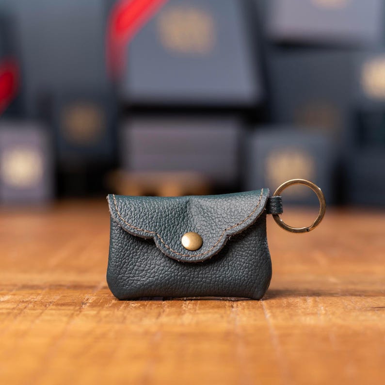 May include: A dark green leather pouch keychain featuring a textured surface, a scalloped flap closure secured with a single brass-colored metal snap button, and a circular brass-colored metal split ring attached to the side for keys.