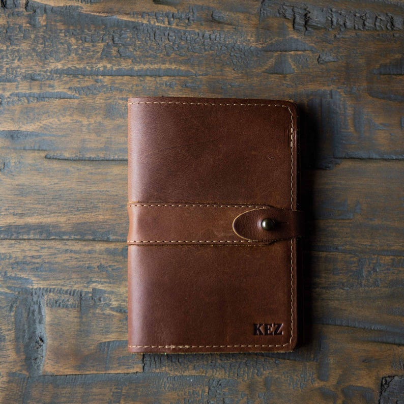 May include: A rectangular brown leather notebook cover featuring a horizontal strap with a single metal snap button closure. The bottom right corner is embossed with the letters KEZ in a simple serif font, and the edges are finished with stitching.