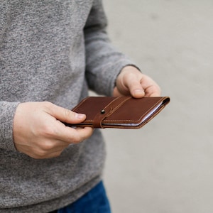 May include: A rectangular brown leather notebook cover featuring a thin strap closure with a small metal stud fastener. The cover has visible stitching along the edges and a smooth, matte texture, designed to hold a slim paper insert securely.