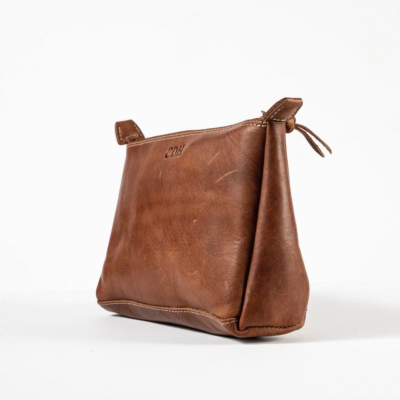 May include: A brown leather pouch with a zippered top closure and visible stitching along the edges. The front surface features the embossed letters CDH. The pouch has a structured, trapezoidal shape with a smooth, matte finish and natural texture.