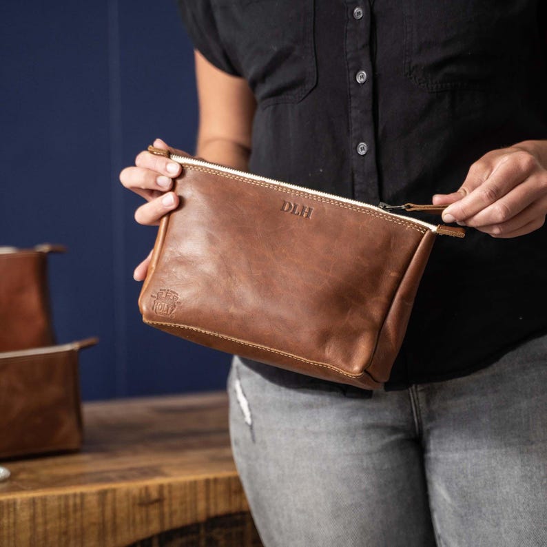 May include: A brown leather zippered pouch featuring the embossed letters DLH in the center and a small circular logo stamp in the bottom left corner. The rectangular accessory has a top zipper closure with a matching leather pull tab.