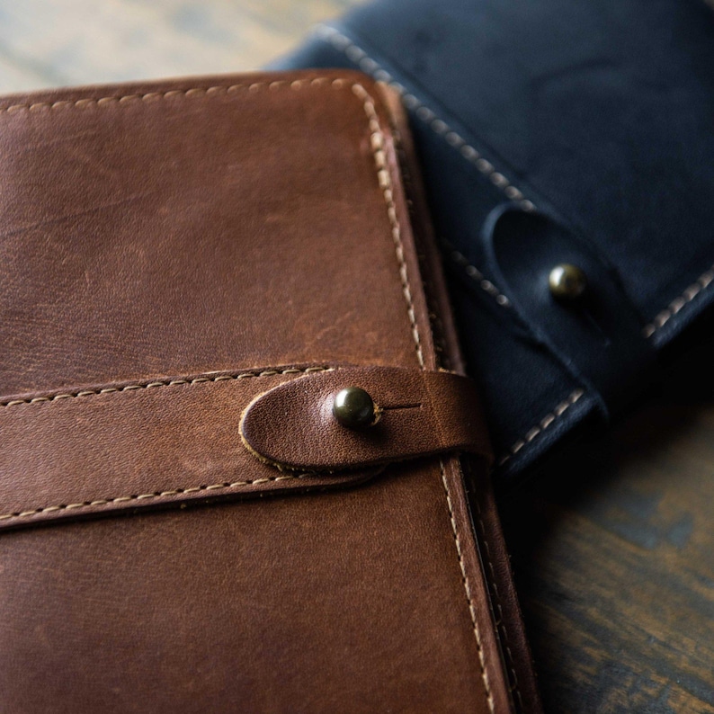 May include: A brown leather journal cover with visible stitching and a strap closure secured by a small brass stud. A second, identical journal cover in black leather is partially visible in the background, featuring the same strap and stud design.
