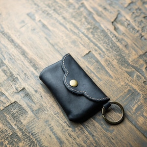 May include: A black leather key pouch featuring a scalloped flap, a brass-toned snap button closure, light-colored contrast stitching along the edges, and a circular brass-toned metal key ring attached to the side of the rectangular case.