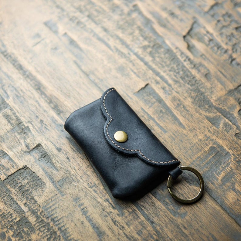 May include: A black leather key pouch featuring a scalloped flap, a brass-toned snap button closure, light-colored contrast stitching along the edges, and a circular brass-toned metal key ring attached to the side of the rectangular case.