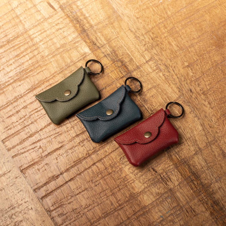 May include: Three small rectangular leather pouches with a pebbled texture, each featuring a scalloped flap, a central brass-colored snap button closure, and a circular metal split ring attached to the side in olive green, navy blue, and red.