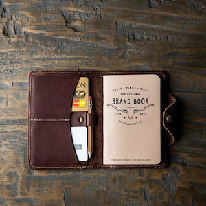 May include: A dark brown leather notebook cover featuring a vertical pocket containing two credit cards and a wooden pencil. The open cover holds a tan paper notebook with the text "THE ORIGINAL BRAND BOOK" and a longhorn steer illustration.