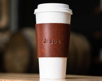 May include: A white disposable coffee cup featuring a brown leather sleeve wrapped around the center. The leather sleeve is embossed with the name Jason in a serif font. The cup has a matching white plastic lid and a smooth, cylindrical shape.