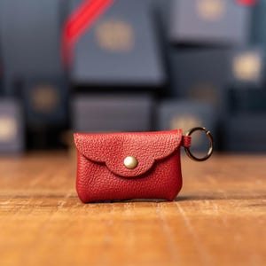 May include: A small red leather pouch with a pebbled texture, featuring a scalloped flap closure secured by a single brass-colored metal snap button. A circular metal key ring is attached to the side of the pouch, providing a functional accessory.