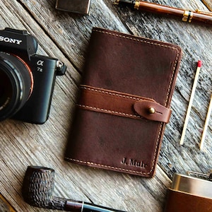 May include: A dark brown leather notebook cover featuring a horizontal strap with a brass stud closure. The leather has visible stitching along the edges and the name J. Muir is embossed in the bottom right corner of the front cover.