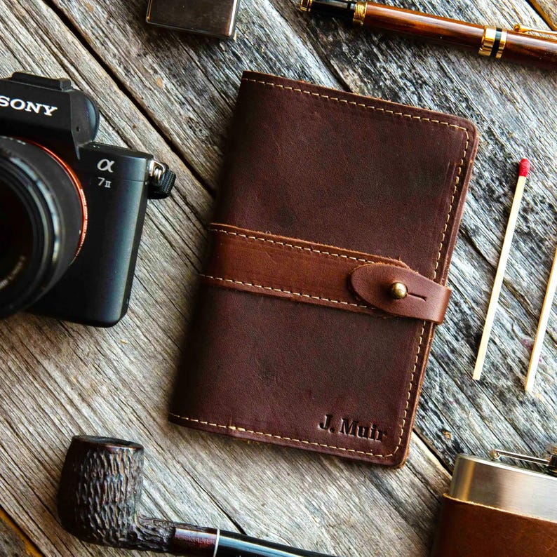 May include: A dark brown leather notebook cover featuring a horizontal strap with a brass stud closure. The leather has visible stitching along the edges and the name J. Muir is embossed in the bottom right corner of the front cover.