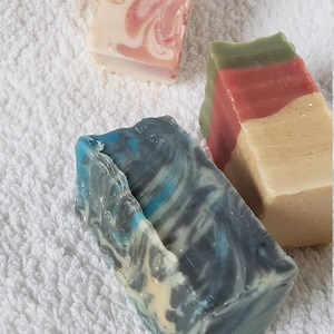 May include: Three bars of handmade soap. One bar is pink and white with swirls, one is green, red, and beige, and one is blue and white with swirls.