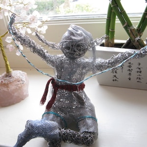 Puede incluir: Una escultura de alambre plateado de una persona con una faja roja y detalles azules. La escultura está en posición de brazos extendidos y sostiene un alambre azul. La escultura está sentada sobre una superficie blanca.