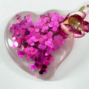 Puede incluir: Un adorno de resina en forma de corazón rosa relleno de purpurina rosa y coronado con una flor rosa y blanca.