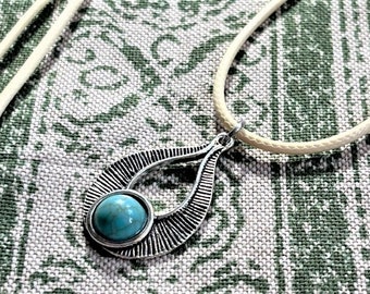 Puede incluir: Un collar con un colgante de piedra turquesa en una montura de plata en forma de lágrima. El colgante tiene un diseño texturizado y está suspendido de un cordón de color crema.
