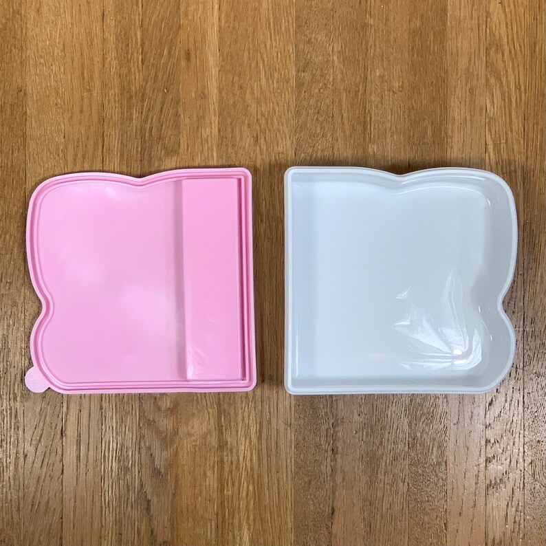 May include: Two plastic containers, one pink and one white, shaped like a book with rounded edges. Both containers are empty.