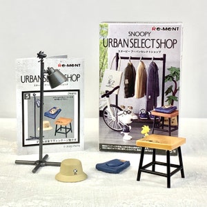 Puede incluir: Un conjunto en miniatura de artículos de tienda urbana con temática de Snoopy, que incluye un perchero con tres chaquetas, un taburete, un sombrero, un par de vaqueros y un soporte de luz. El conjunto forma parte de la colección Re-Ment Snoopy Urban Select Shop.