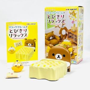 May include: A Rilakkuma room miniature set featuring a brown bear bed, a yellow duck figure, and a box with Japanese text. The bed has a yellow blanket with white polka dots. The box is yellow and has Japanese text and illustrations.