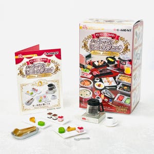 May include: Miniature food set with a box and booklet. The box features images of various dishes, while the booklet shows miniature desserts. Several miniature food items, including cakes and pastries, are arranged on small plates.