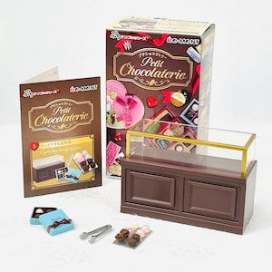 May include: A miniature chocolate shop display set. The set includes a brown display case with a glass top, boxes, and miniature chocolates. The packaging features the text "Petit Chocolaterie" and Japanese text. The set also includes a small pair of tongs.